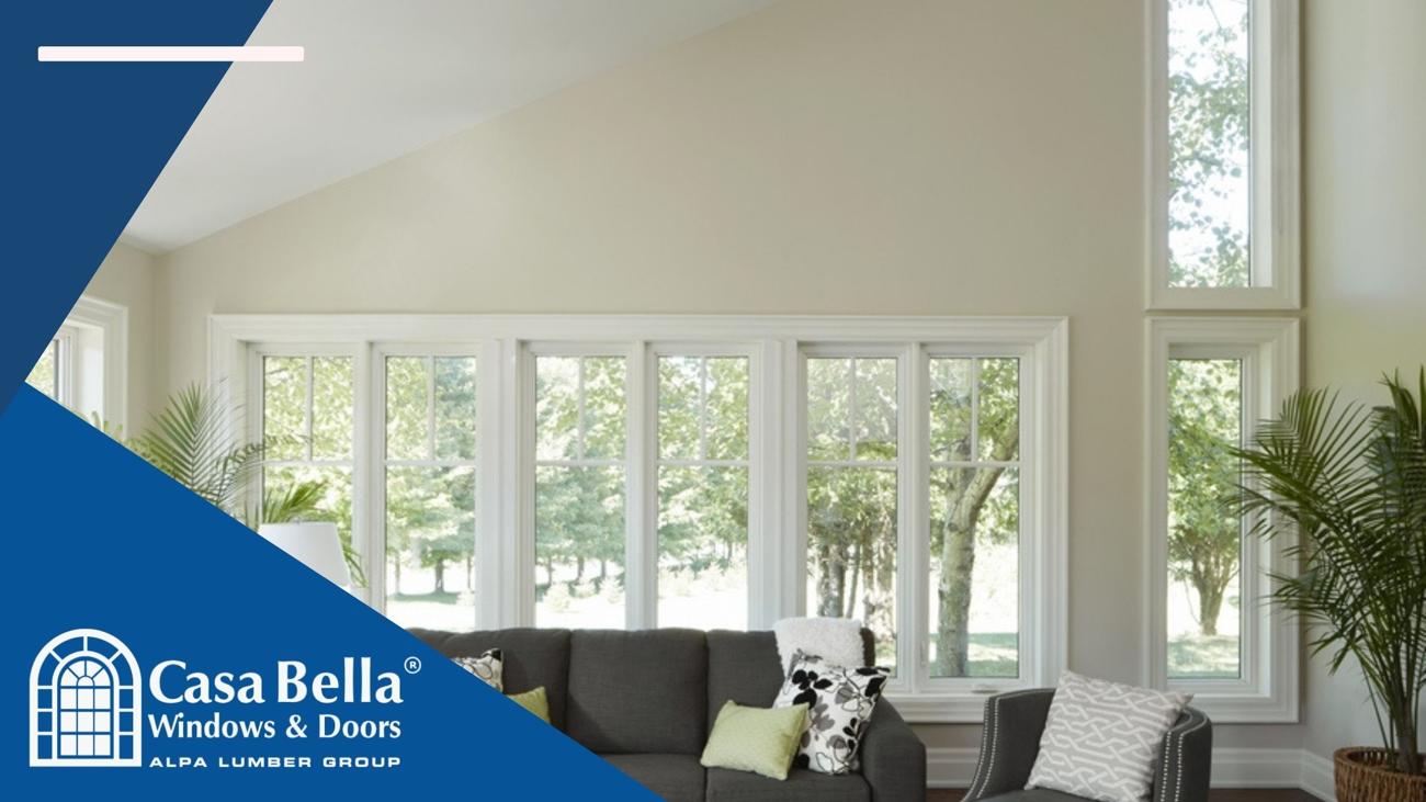 A bright and modern living room featuring large, elegant windows with white trim, allowing natural light to enter. The Casa Bella Windows & Doors logo is displayed in the bottom left corner.