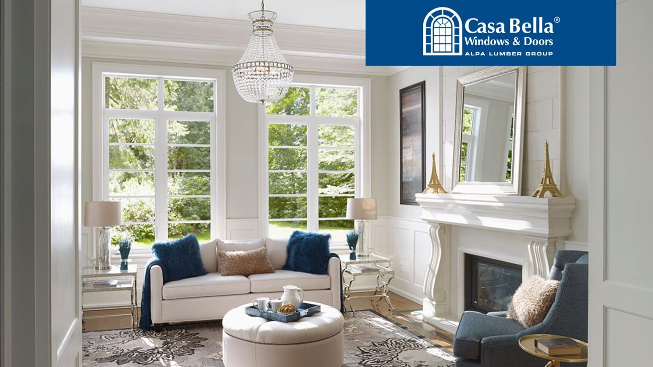 A bright, elegant living room with large windows allowing natural light to fill the space. The room features a white sofa, plush decor, a fireplace with a mirror, and a chandelier. The Casa Bella Windows & Doors logo is displayed in the top right corner.