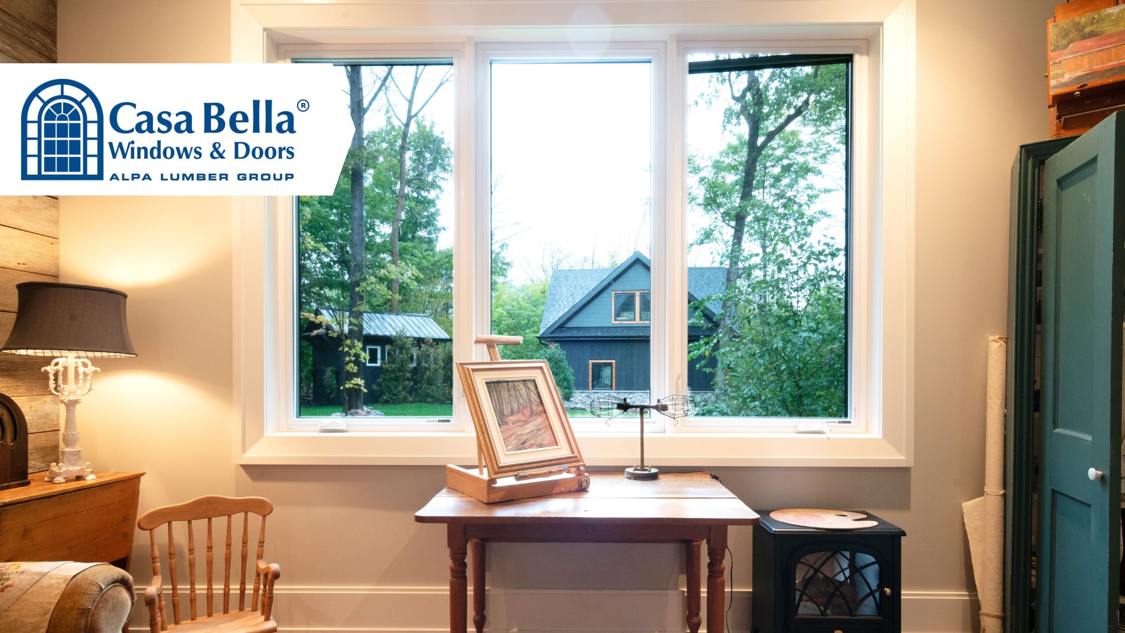 Interior view of a cozy living space with Casa Bella’s large picture window, offering a scenic view of a cottage and trees, and bringing abundant natural light into the room.